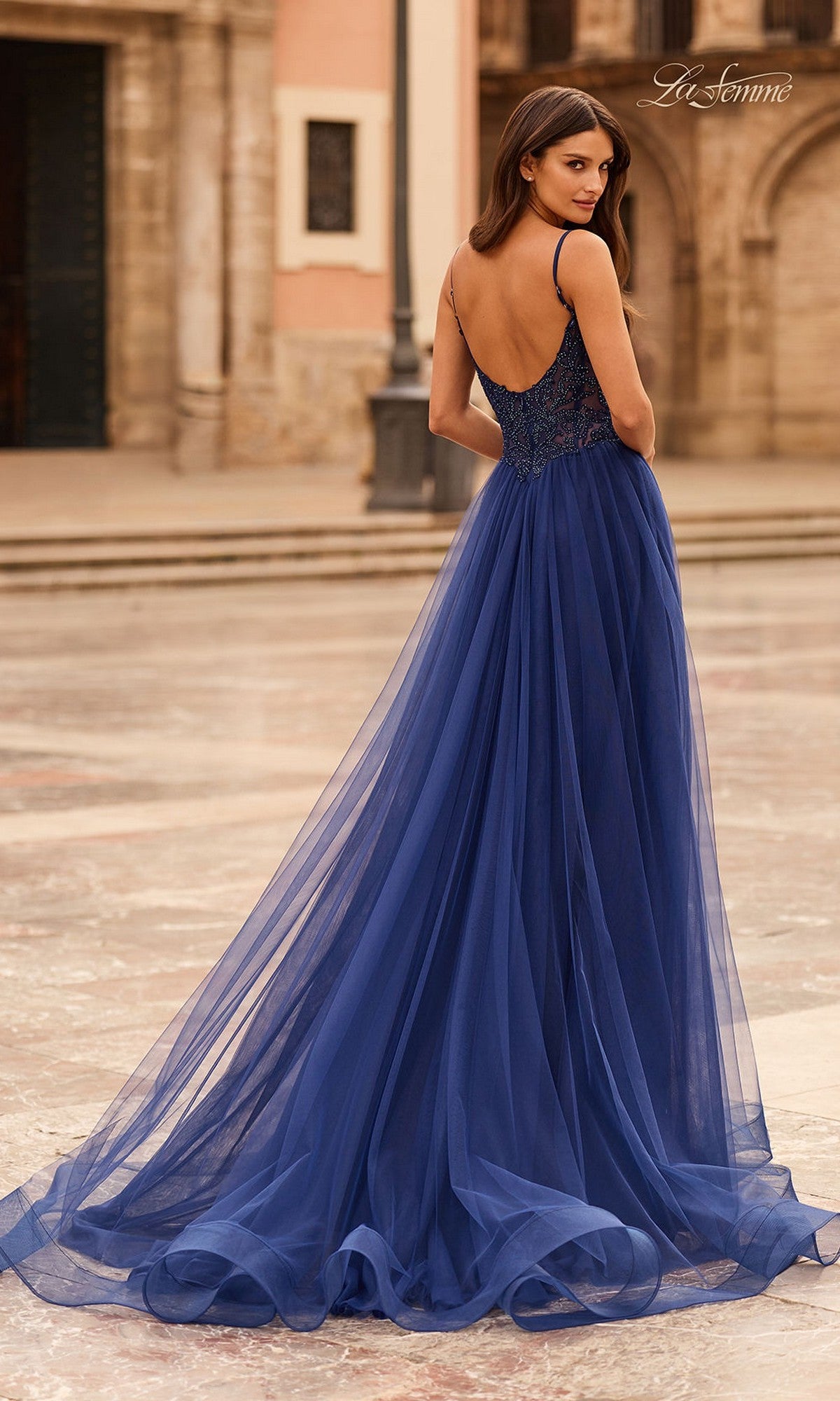 Wearing the La Femme 33674 long prom dress, a woman stands outdoors on marble in a sleeveless, floor-length deep blue gown with a beaded corset bodice and low back, glancing over her shoulder.